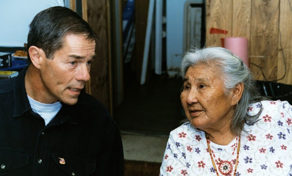 A middle-aged white man and Alaska Native elder woman sitting, leaning in, and talking to each other.