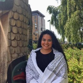 A photo of Rellani Ogumoro wrapped in a traditional shawl by a totem pole