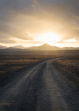 Photo of dirt road leading to sunlit mountains in distance