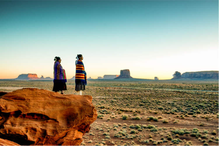 Two Native American people wrapped in striped blankets looking at the sunset from a red rock across the high desert