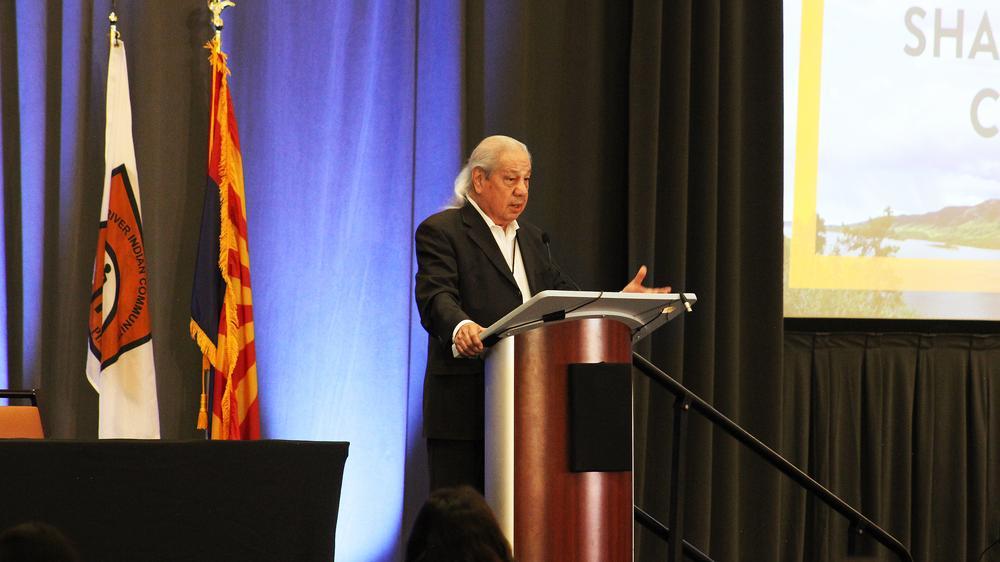 Older man with long white hair speaking at lectern. 